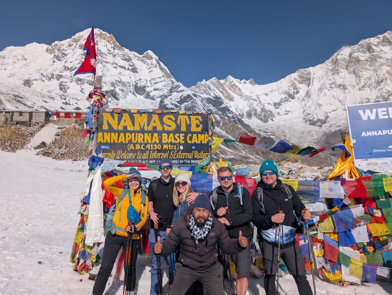 A group of six trekkers stands smiling in front of a sign that reads "NAMASATE ANNAPURNA BASE CAMP" with prayer flags and snowy mountain scenery in the background.